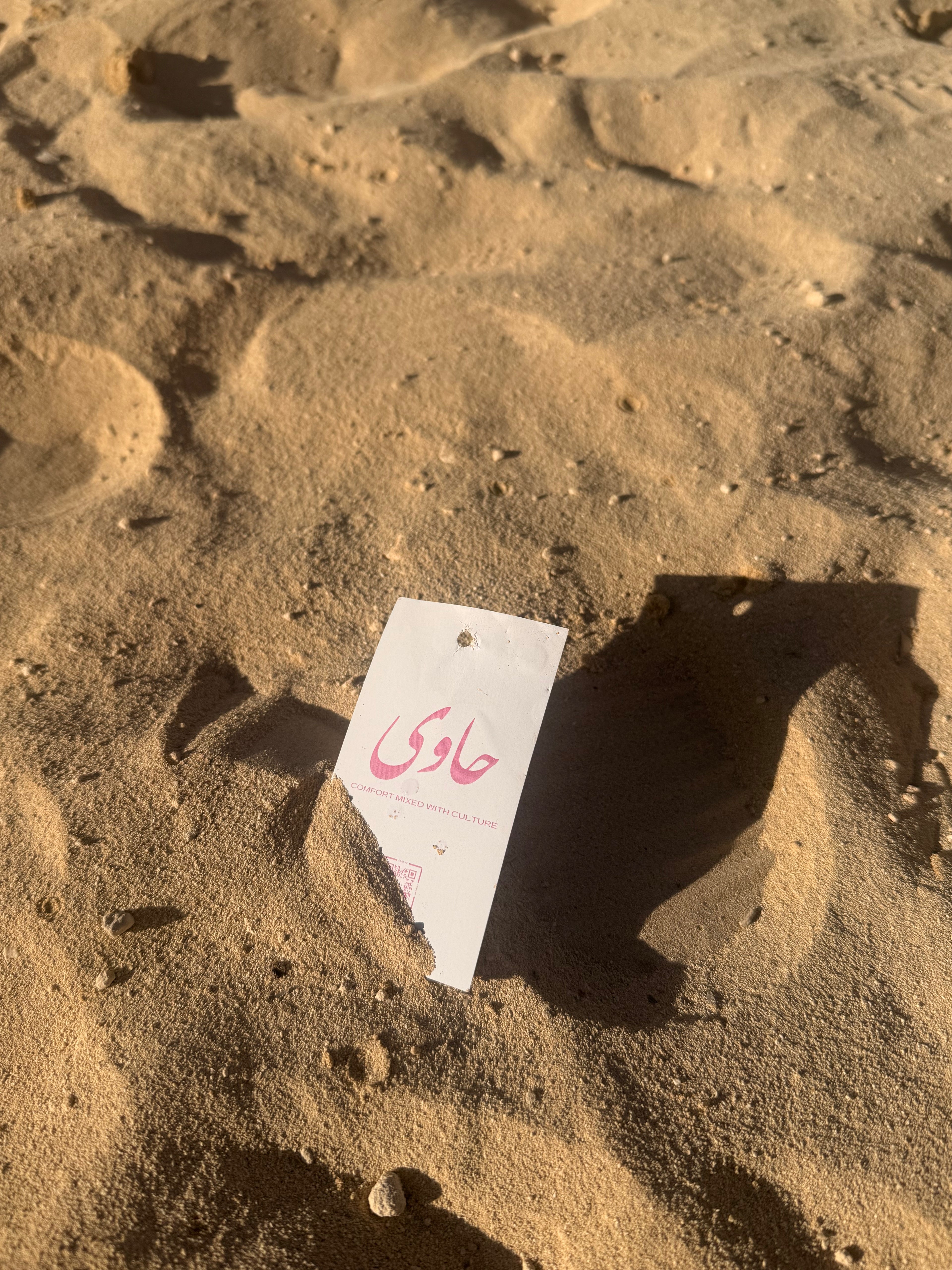 A small white paper tag with pink Arabic text lies on sandy ground outdoors, casting a strong shadow in the sunlit sand.
